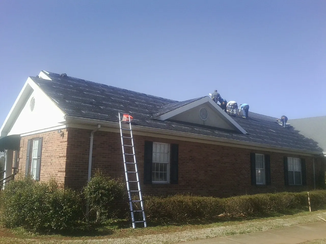 Crew tearing shingles off a brick ranch roof with a ladder set — Emergency Roof Repair work by our crew in Honeoye Falls