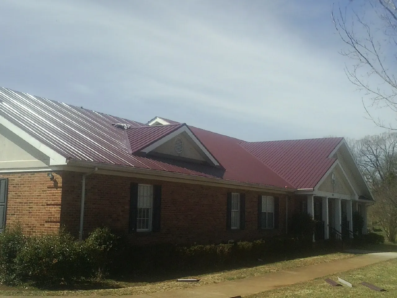 Red metal roof with gabled entries on a long brick building, from a Hail Damage Roof Repair job in Honeoye Falls
