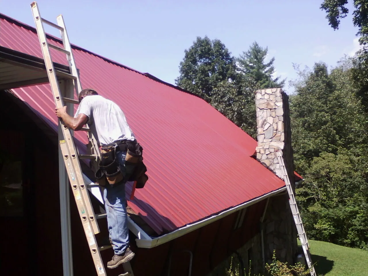 Roofer on a ladder beside a red metal roof and stone chimney — Soffit & Fascia Repair by Roofing Brothers in Honeoye Falls